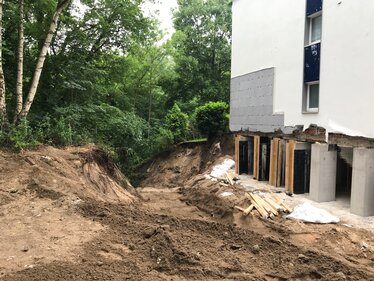 Freigelegte Gebäudefundamente und unbefestigter Boden entlang des Wohngebäudes nach Unterspülung. Die Aufnahme zeigt den sanierten Bereich mit sichtbaren Stützmaßnahmen zur Sicherung des Gebäudes und des angrenzenden Geländes.
