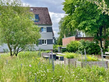 Blütenreiche Grünfläche mit Hochbeeten und Sitzgelegenheiten vor einem Wohngebäude. Die naturnah gestaltete Außenanlage bietet Lebensraum für Insekten und Aufenthaltsmöglichkeiten für Anwohnende.
