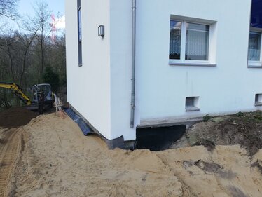 Sanierungsarbeiten an dem Wohngebäude nach Unterspülung. Freigelegte Gebäudefundamente werden mit Erdarbeiten und Abdichtungsmaßnahmen gesichert.