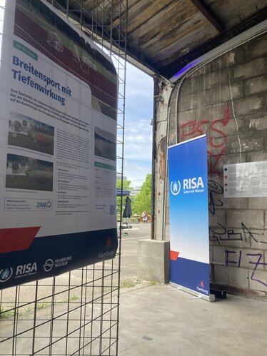 Das Bild zeigt einen Aufsteller mit dem RISA-Logo sowie ein Plakat mit Informationen zum Hein-Klink-Stadion, welches bei Starkregen Regenwasser zur Entlastung des Sielsystems aufnehmen kann