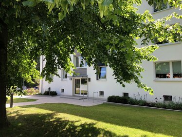 Eingangsbereich eines Mehrfamilienhauses mit gepflegter Grünanlage und schattenspendendem Baum. Vor dem Gebäude befinden sich Rasenflächen, Beete mit Stauden sowie befestigte Wege.
